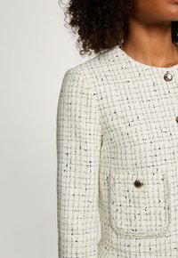 Giacca in tweed bianco con motivo a reticolo, caratterizzata da un collo rotondo, bottone dorato e tasca frontale. Tessuto testurizzato con accenti neri.