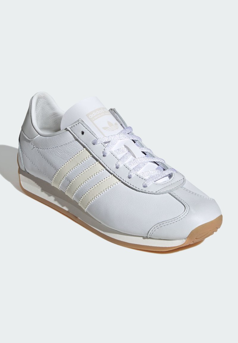 Zapatilla deportiva de piel blanca con tres franjas color crema, detalles grises, puntera redondeada, lengüeta texturizada y suela de goma. Cordones reflectantes.