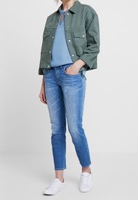 Olivgrön jacka med två framfickor, blå topp, ljusblå jeans och vita sneakers. Avslappnat outfit med en avslappnad passform.