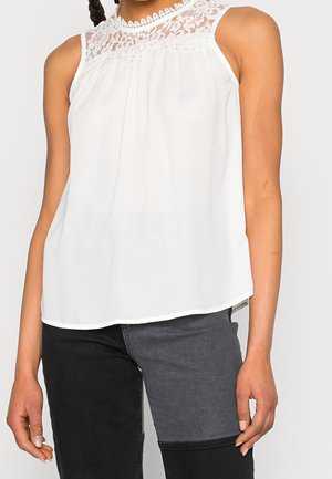 Ærmeløs hvid bluse med blonde detaljer på skuldrene og den øverste del af brystet, kombineret med sort og grå to-farvet jeans.