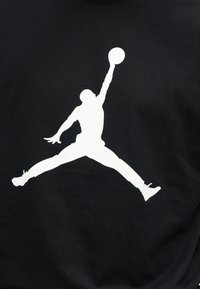 Σιλουέτα παίκτη μπάσκετ που πηδά με τα πόδια ανοιχτά, εκτείνει το ένα χέρι προς τα πάνω κρατώντας μια μπάλα μπάσκετ σε μαύρο φόντο.