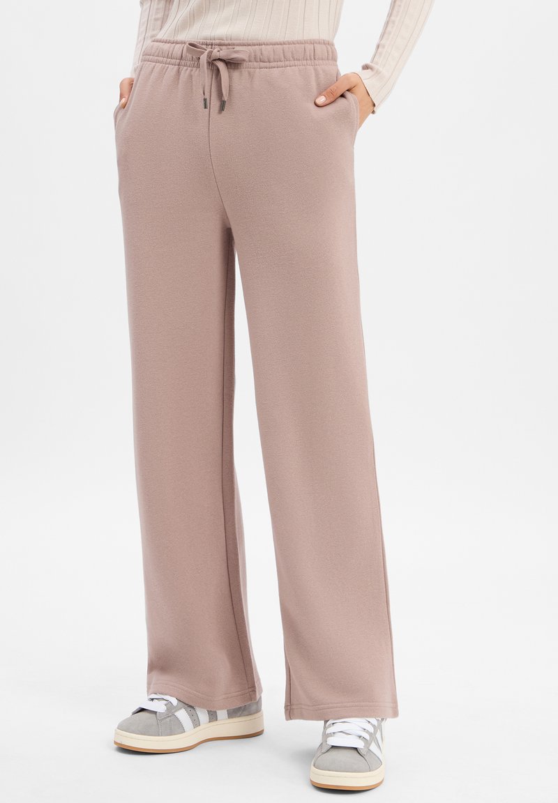 Kobieta w wysokich, szerokich spodniach w odcieniu mauve z troczkami, z rękami w kieszeniach, w połączeniu z beżowymi sneakersami i dzianinowym swetrem w kremowym kolorze.