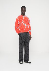 Κόκκινο φούτερ με floral σχέδιο και μπλε λεπτομέρειες, συνδυασμένο με μαύρο παντελόνι που έχει έντονο λουλουδένιο σχέδιο. Το μοντέλο στέκεται με τα χέρια στις τσέπες.