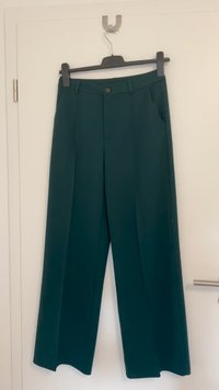 Groene, wijde broek van gladde stof. Voorzien van een knoopsluiting, riemlussen en zijzakken. Hangt aan een zwarte hanger.