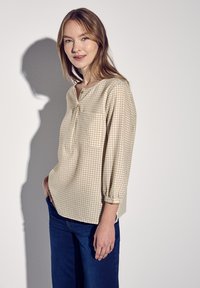 Blusa beige a quadri con scollatura a V, maniche a tre quarti e taschino sul petto. Indossata con pantaloni in denim blu su uno sfondo chiaro.