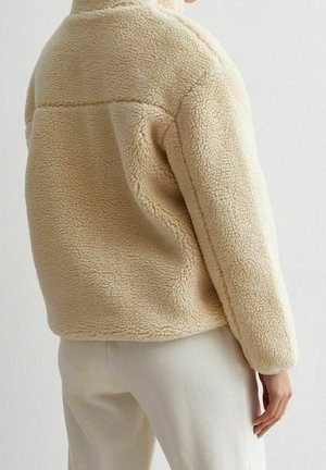 Osoba nosząca beżową, teksturowaną kurtkę polarową oraz jasne spodnie, widziana od tyłu na jednolitym tle.