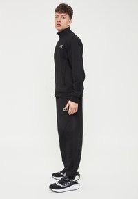 Felpa nera con zip e colletto alto, abbinata a pantaloni neri coordinati e sneakers nere con dettagli bianchi.