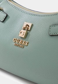 Miętowa torebka ze skóry o fakturze, wyposażona w złote okucia i tabliczkę z logo "GUESS EST 1981". Zamek błyskawiczny i widoczne elementy ozdobne.