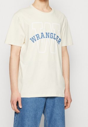 T-shirt en coton couleur crème avec un grand texte « WRANGLER » en bleu sur le devant, coupe décontractée et manches courtes.