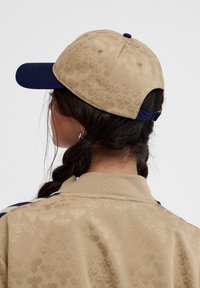 Tanfarbene Baseballmütze mit einem marineblauen Schild, ausgestattet mit einem tonal geprägten Muster. Verstellbarer Riemen hinten für eine individuelle Passform.