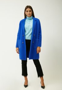 Helles blauer Wollmantel mit zwei vorderen Taschen und schwarzen Knöpfen, getragen über einem hellblauen Rollkragenpullover und schwarzen Jeans mit Spitze.