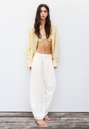 Tânără cu păr lung și închis la culoare poartă o cămașă deschisă galbenă cu dungi peste un sutien de bikini galben, pantaloni albi cu picioare largi și sandale bej.