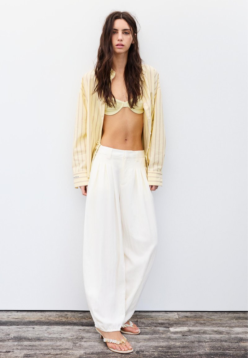 Jeune femme aux longs cheveux foncés porte une chemise ouverte à rayures jaunes sur un haut de bikini jaune, un pantalon blanc à jambes larges et des sandales beiges.