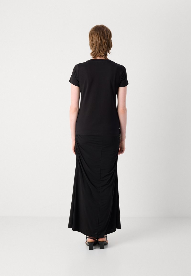 Femme debout, de dos, portant un haut noir à manches courtes, une longue jupe noire et des sandales à talons noires sur un fond uni.