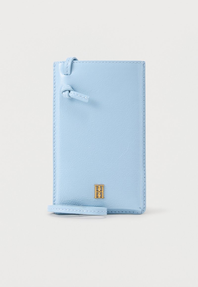 Δερμάτινο θήκη τηλεφώνου ανοιχτού γαλάζιου χρώματος με λουράκι καρπού και μικρό χρυσό έμβλημα στο μπροστινό μέρος, σε απλό λευκό φόντο.