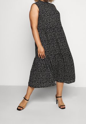 Vrouw draagt een zwarte mouwloze midi-jurk met witte stippen en zwarte sandalen met hoge hakken, staand tegen een effen achtergrond.