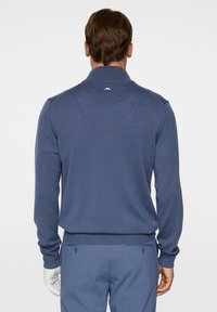 Miestsky tmavomodrý pletený sveter s golierom a dlhými rukávmi, s rebrovanými manžetami a spodným okrajom. Detail loga na zadnej časti výstrihu.
