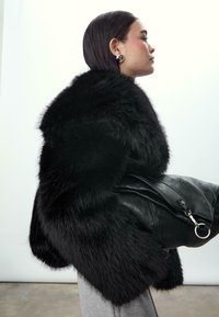 Cappotto di pelliccia nera con un colletto voluminoso, abbinato a una borsa in pelle nera con dettagli metallici e una superficie testurizzata.