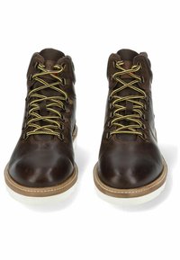 Hnědé kožené turistické boty se žlutými tkaničkami, kovovými očky, bílou gumovou podrážkou a texturovaným povrchem. Středně vysoký design pro podporu kotníku.