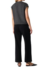 Frau in einem ärmellosen, dunkelgrauen Stricktop, schwarzen geraden Hosen und schwarzen Slingback-Flats mit spitzer Zehe, von hinten gesehen.