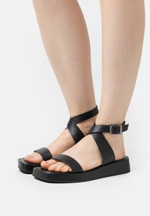 Sandales en cuir noir avec des brides croisées et une sangle de cheville dotée d'une boucle. Semelle plate avec une finition texturée, design à bout ouvert.