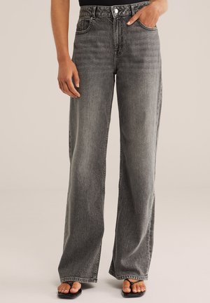 Femeie purtând blugi negri depăliți, cu talie înaltă și croială largă, cu nasturi și fermoar, asortați cu sandale negre tip thong, pe un fundal neutru.