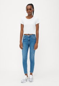 Camiseta blanca de manga corta con borde ondulado, combinada con jeans ajustados azules con detalles desgastados y zapatillas blancas con acentos grises.