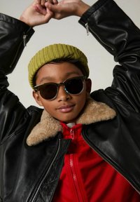 Veste en cuir noir avec col en sherpa, sweat à capuche rouge zippé, bonnet en maille vert, et lunettes de soleil rondes avec verres sombres.