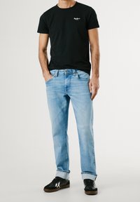 Homme portant un t-shirt noir à manches courtes, un jean bleu clair avec des revers et des baskets noires, debout avec une main dans la poche.