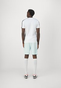 Camiseta deportiva blanca con franjas laterales negras, combinada con pantalones cortos de color azul claro y calcetas blancas hasta la rodilla. Botas de fútbol negras con acentos neón.