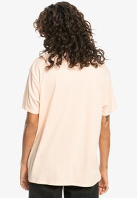 Lyhythihainen, vaalean persikan värinen t-paita, joka on valmistettu pehmeästä puuvillasta. Rentoa mitoitusta, pyöreä pääntie, takana yksinkertainen muotoilu ilman näkyviä logoja tai kuvioita.