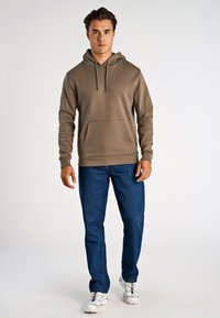 Sudadera con capucha marrón y bolsillo frontal, combinada con jeans azul oscuro y zapatillas blancas con acentos multicolor. Textura suave.