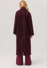 Długi, oversize płaszcz w ciemnym burgundzie, wykonany z teksturowanej, puszystej tkaniny. Posiada duży kołnierz oraz boczne slitsy dla swobody ruchów.