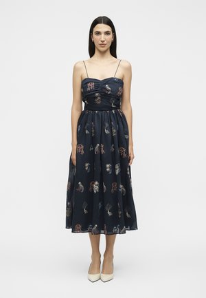 Femme debout portant une robe midi bleu marine à fines bretelles avec des imprimés abstraits multicolores, associée à des talons blancs pointus.