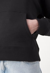Sweat-shirt noir avec une poche kangourou, poignets et ourlet côtelés ; fabriqué en tissu doux, corps partiellement visible en denim clair.
