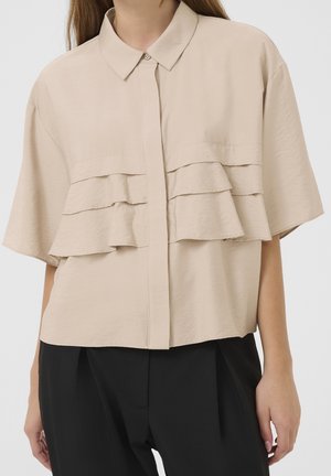 Kobieta w beżowej bluzce z krótkim rękawem z falbanami oraz czarne spodnie z wysokim stanem, stojąca na jednolitym tle.