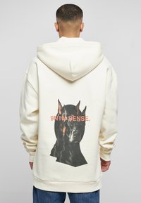 Krém színű kapucnis pulóver, hátul egy nagy fekete és narancssárga doberman grafikával, rajta a "9N1M SENSE" szöveggel. Texturált anyag, laza illeszkedés.