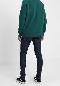 Tmavě modré slim-fit džíny s hladkou texturou, ve spojení se zelenou mikinou a černými teniskami s bílou podrážkou.