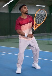 Idrettsutøver i en skjorte med maroon og fersken gradient og korte ermer, kremfargede bukser og hvite sneakers, som holder et lyst oransje tennisracket.
