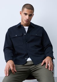 Jeune homme aux cheveux courts portant une veste noire boutonnée sur une chemise blanche et un pantalon olive, assis les mains posées sur les cuisses.