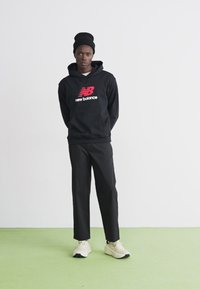 Schwarzer Kapuzenpullover mit rotem und weißem "New Balance"-Logo, kombiniert mit schwarzen Hosen und hellen Sneakers auf einem grünen Boden.