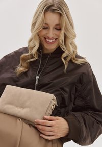 Beige Wildleder-Clutch mit goldenem Logo, vor einem Modell gehalten, das eine schwarze Bomberjacke und ein hellbraunes Outfit trägt.