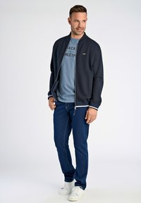 Giacca bomber navy con polsini a costine e zip, indossata sopra una t-shirt azzurro chiaro con testo nero. Jeans in denim e sneakers bianche completano il look.