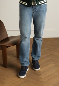 Jeans in denim blu chiaro con taglio dritto, indossati con scarpe in camoscio navy con suole bianche, su un pavimento e una sedia in legno.