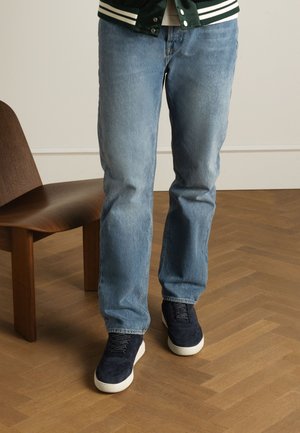 Lichtblauwe denim jeans met een rechte snit, gedragen met marineblauwe suède schoenen met witte zolen, tegen een houten vloer en stoel.