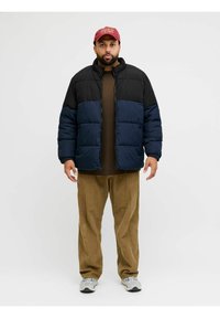 Gepolsterte Jacke mit schwarzem Oberteil und dunkelblauem Unterteil, Reißverschluss und hohem Kragen. Kombiniert mit brauner Cordhose und Turnschuhen.