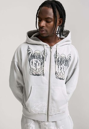 Man met gevlochten haar die naar beneden kijkt en een lichtgrijze hoodie met rits draagt met een schedel en vlammen grafische print en bijpassende broek.