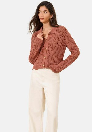 Žena oblečená v tehlovočervenom kardigáne s otvoreným pletením a zlatými gombíkmi a krémových širokých nohaviciach s vysokým pásom, pózujúca s jednou rukou pri golieri.