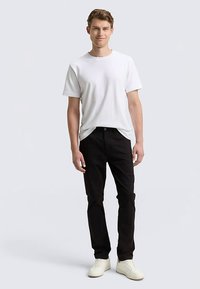 Valge puuvillane T-särk mustade teksadega. Modell seisab valgetes tossudes, esitades juhuslikku sobivust ja neutraalset värvipaletti.