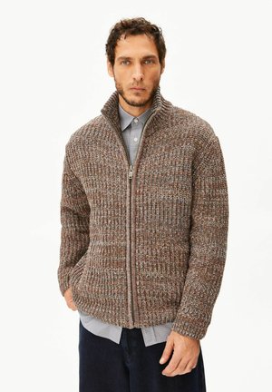 Mann trägt einen braunen und grauen grob gestrickten Reißverschluss-Pullover über einem blau karierten Hemd mit dunkler Hose, steht vor weißem Hintergrund.
